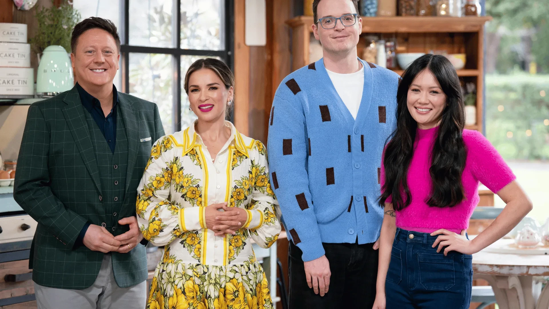 Four presenters standing in a bright kitchen studio on a baking competition show set with ingredients and shelves in the background
