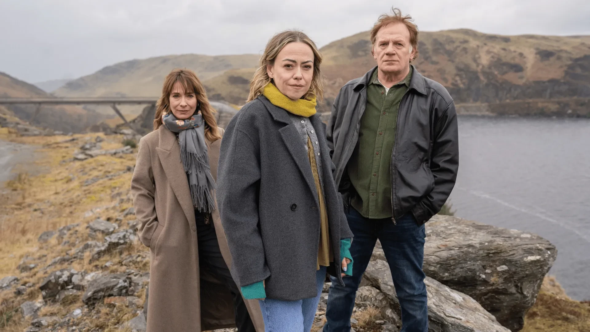 Three people standing on a rocky coastal landscape overlooking water and hills, moody crime drama setting