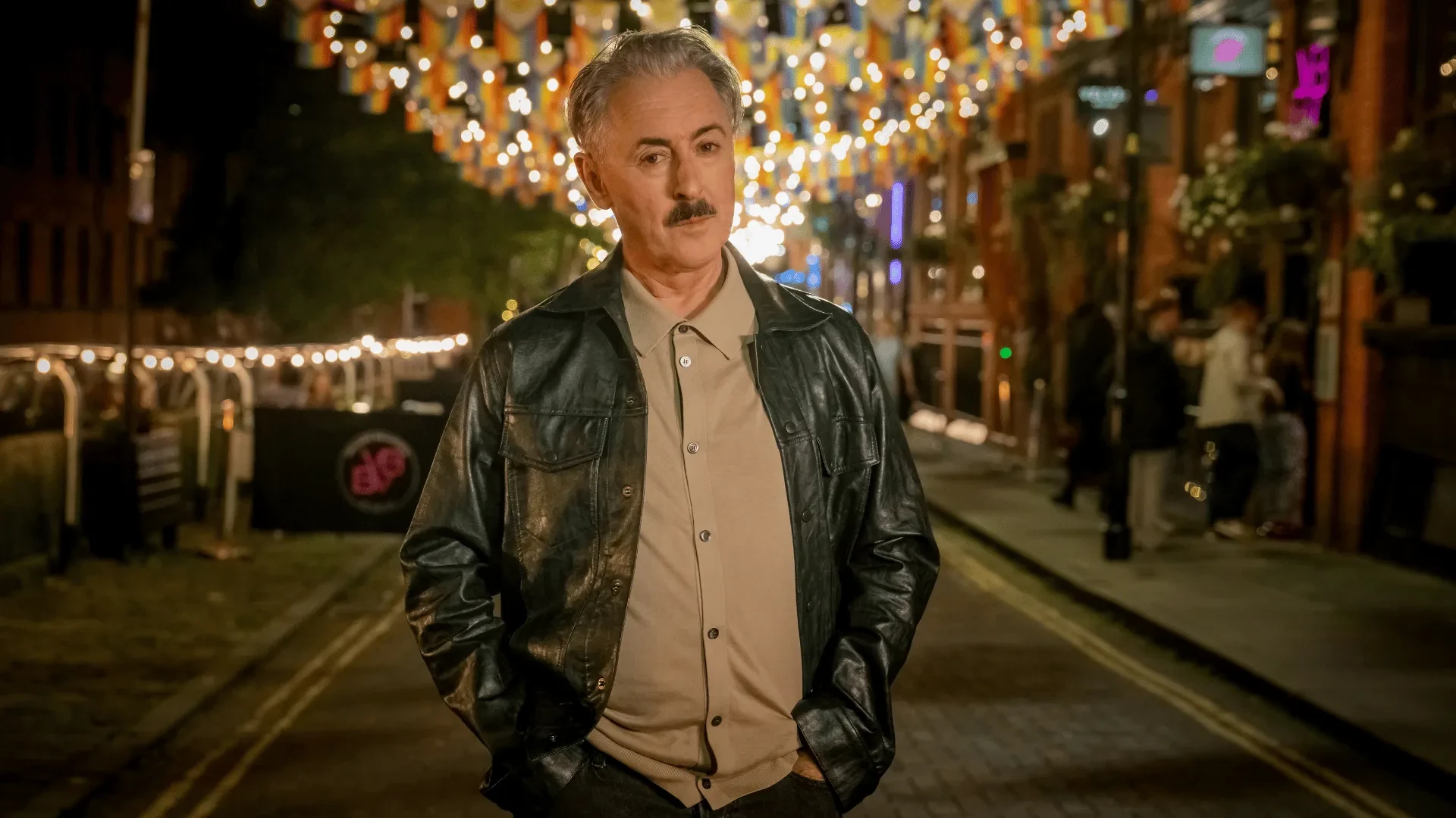 Man in black leather jacket standing on a city street at night with festive lights and blurred urban background