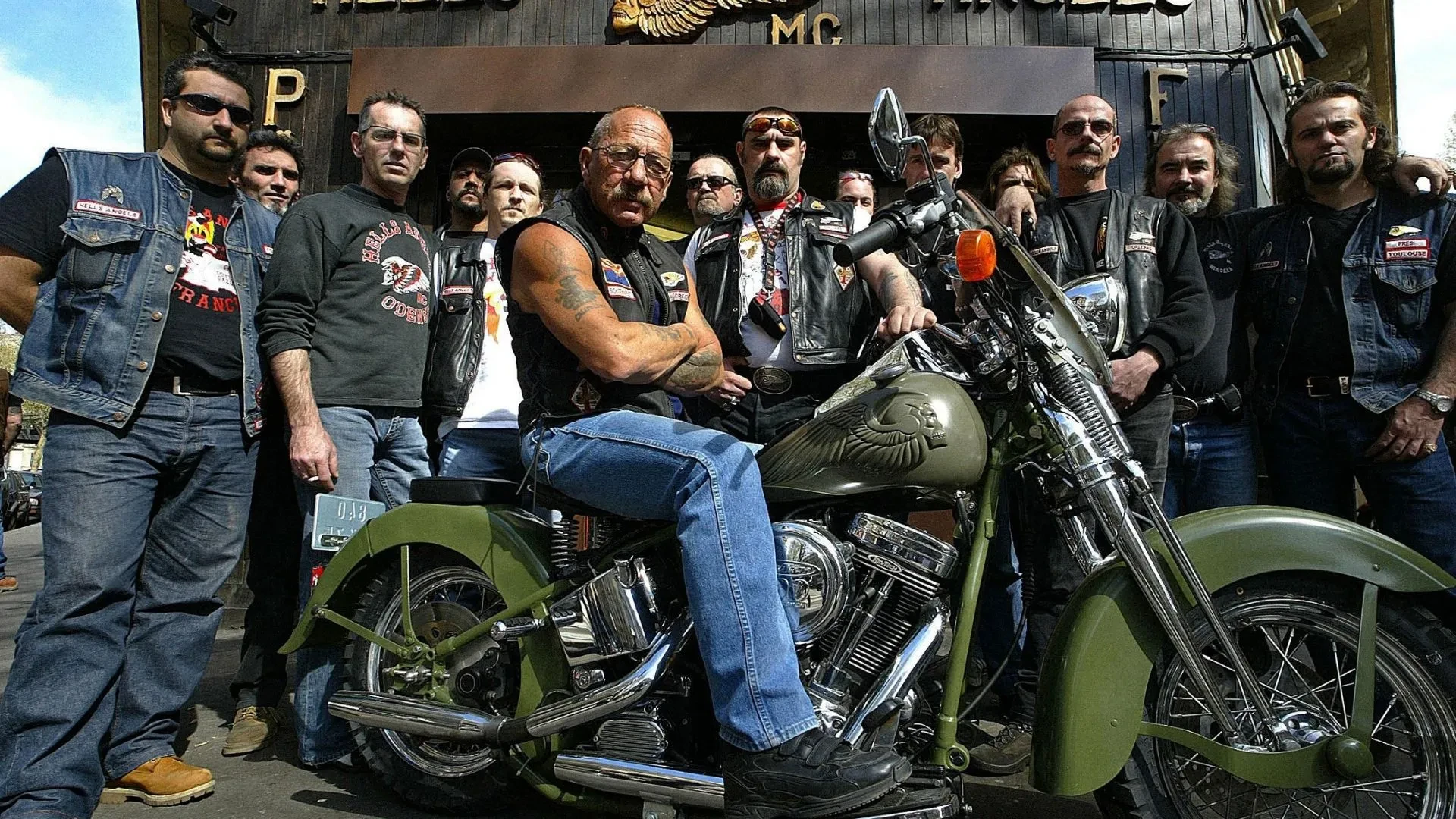 Members of a motorcycle club gathered around a custom cruiser motorbike outside a clubhouse, showcasing biker culture and motorcycle club community.