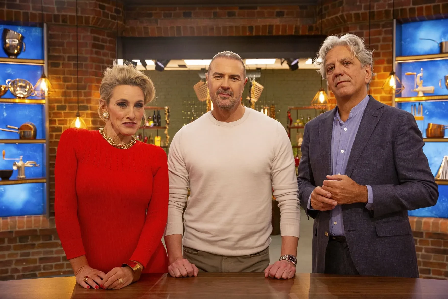 Judges Grace Dent, Paddy McGuinness, and Giorgio Locatelli pose together on the set of MasterChef behind a wooden table