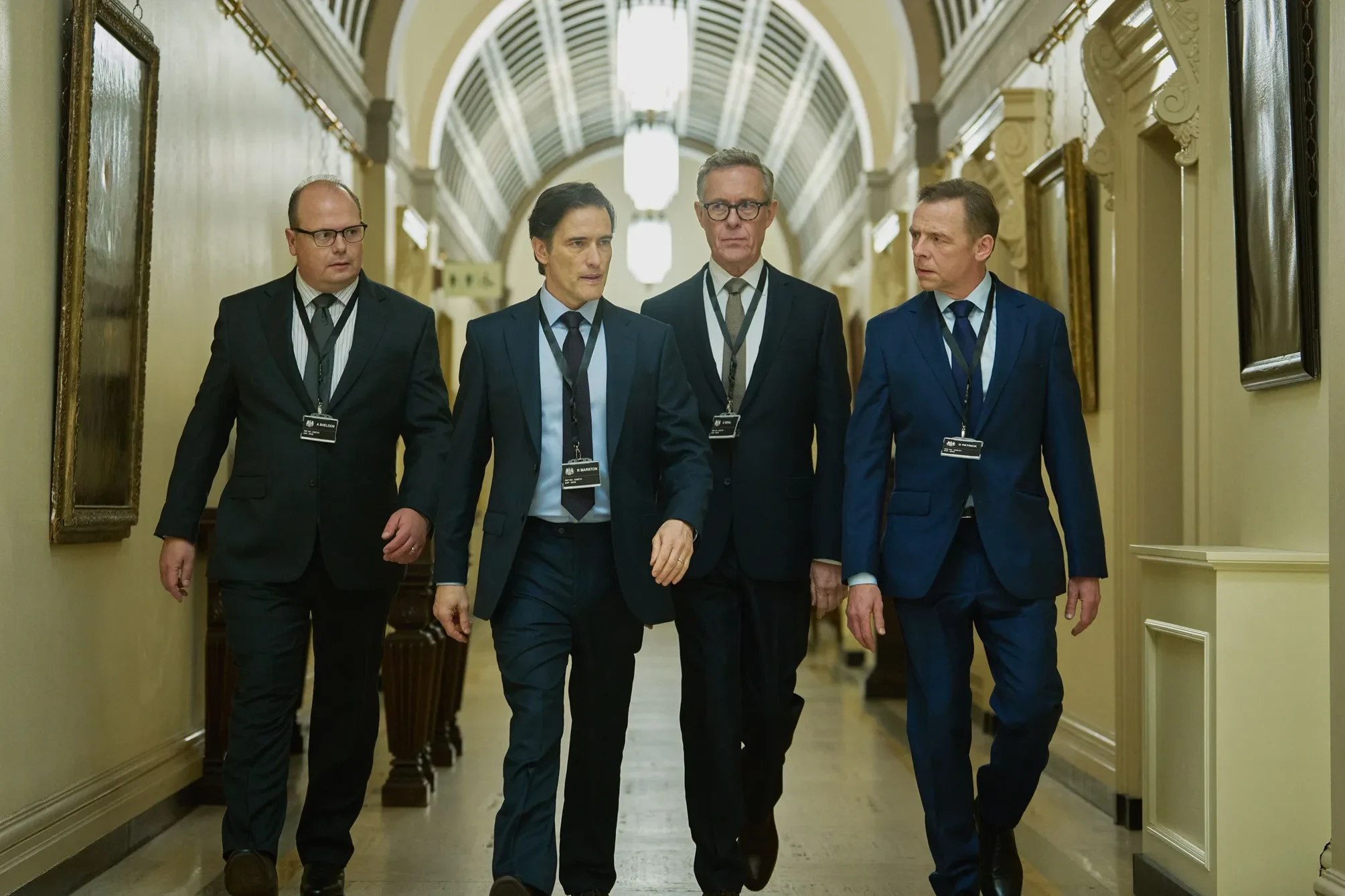 Four men in business suits wearing official lanyard passes walk down a grand government hallway in a still from the drama series The Undeclared War