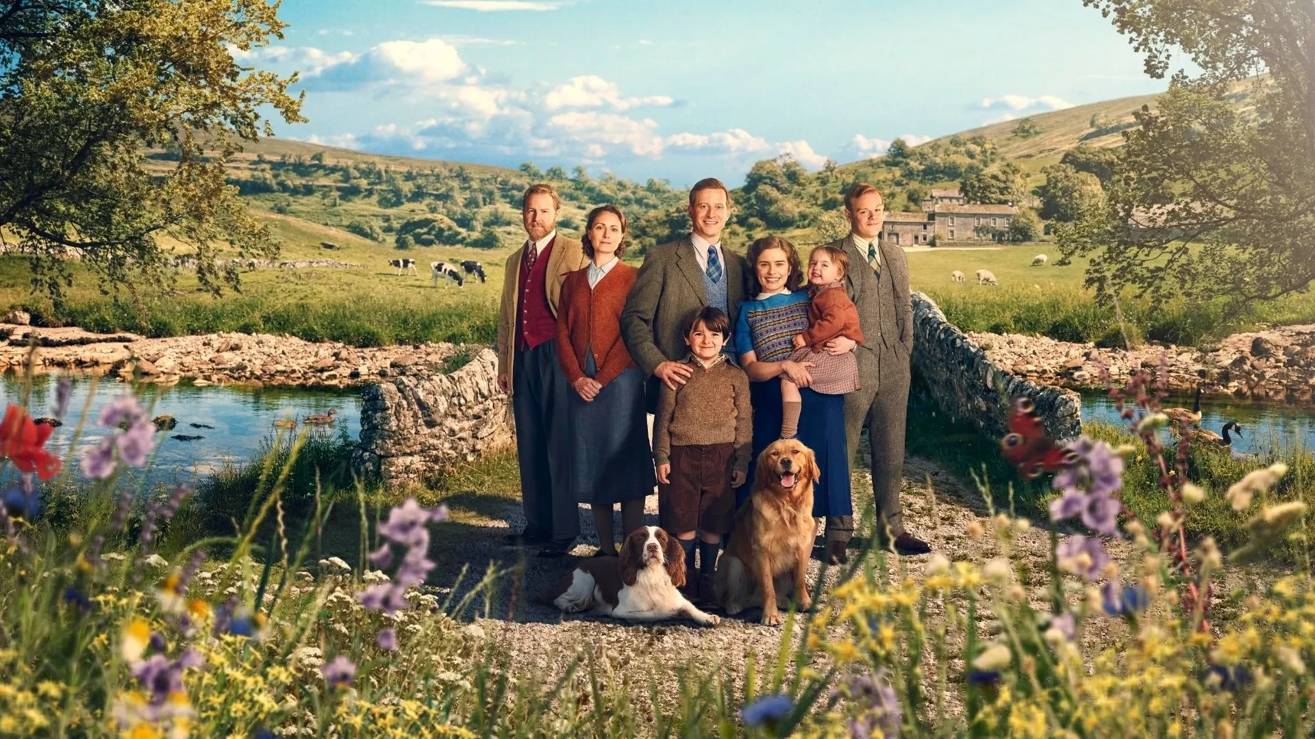 Promotional image for All Creatures Great and Small featuring a group of characters, children and dogs standing on a stone bridge in the Yorkshire countryside surrounded by wildflowers and rolling hills.