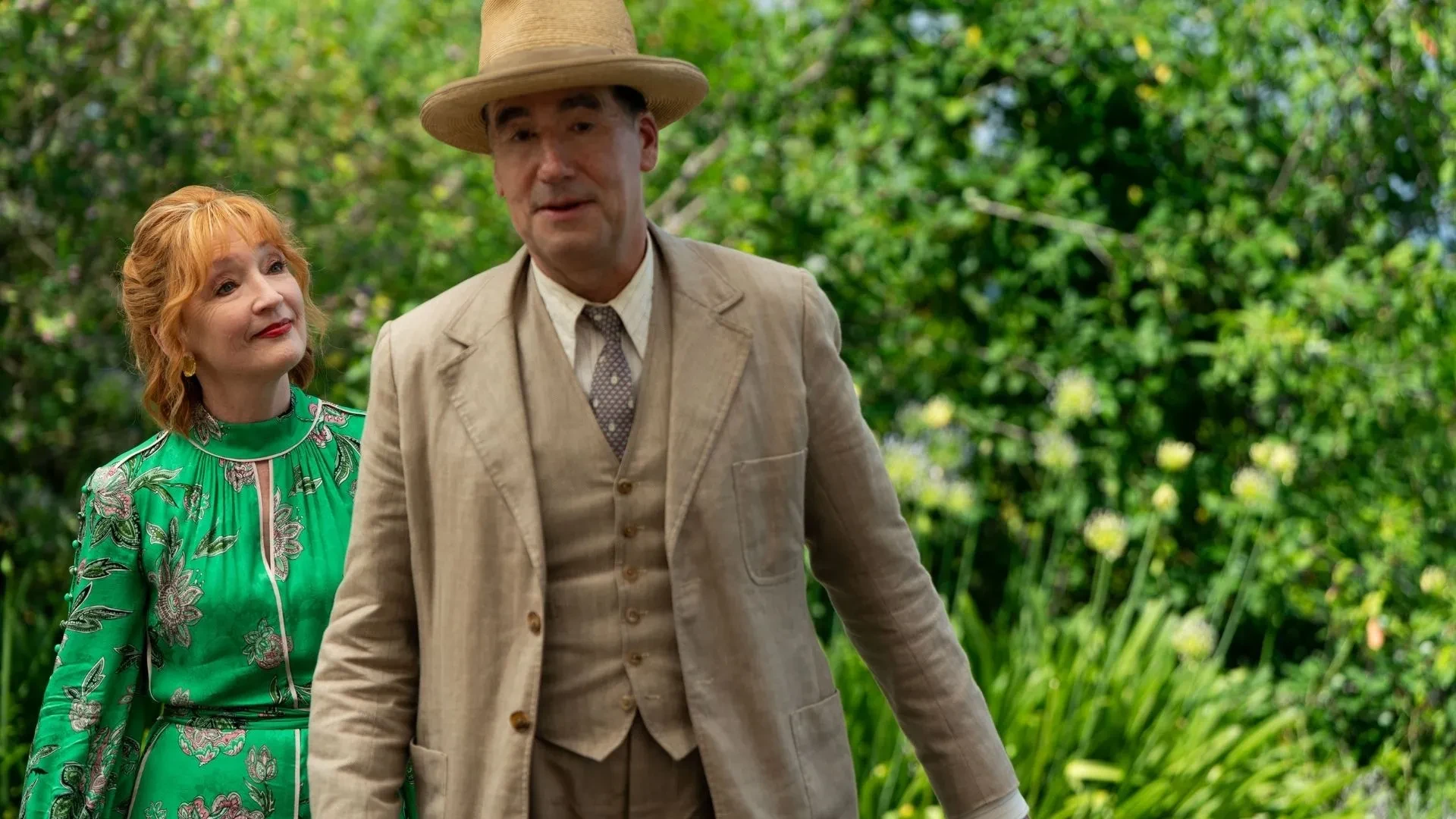 Promotional image for Marble Hall Murders featuring two characters in period clothing standing in a garden surrounded by greenery.