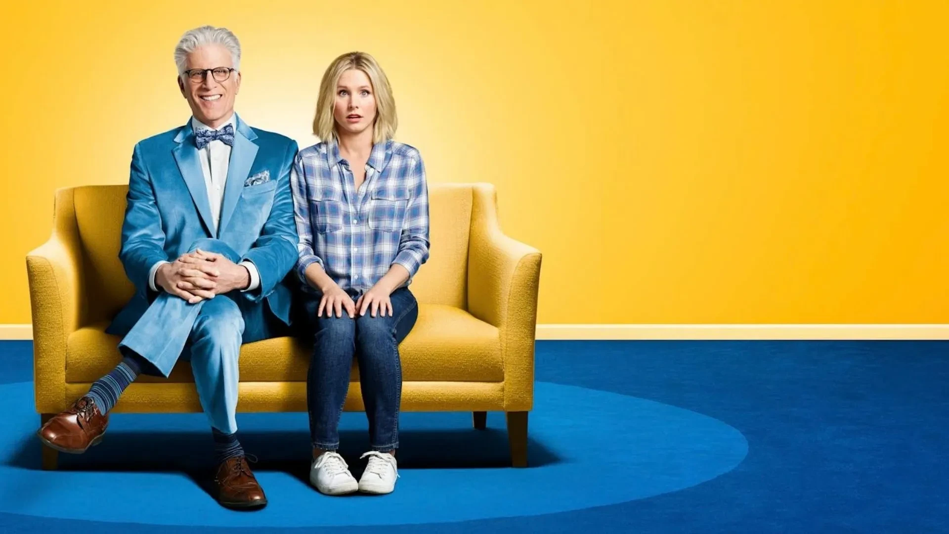 A man and a women wearing blue sat on a yellow sofa on blue carpet against a yellow background. Key art image for The Good Place.